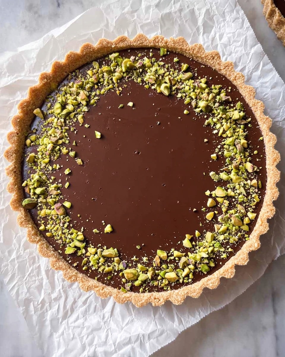 A round tart with three visible layers: the bottom layer is a light brown crumbly crust with scalloped edges, the middle layer is a smooth, glossy dark brown chocolate filling that fills almost all the tart, and the top layer is scattered with small green chopped pistachios mainly around the edge, leaving the center free. The tart rests on crumpled white parchment paper, which is set on a white marbled surface. Photo taken with an iphone --ar 4:5 --v 7