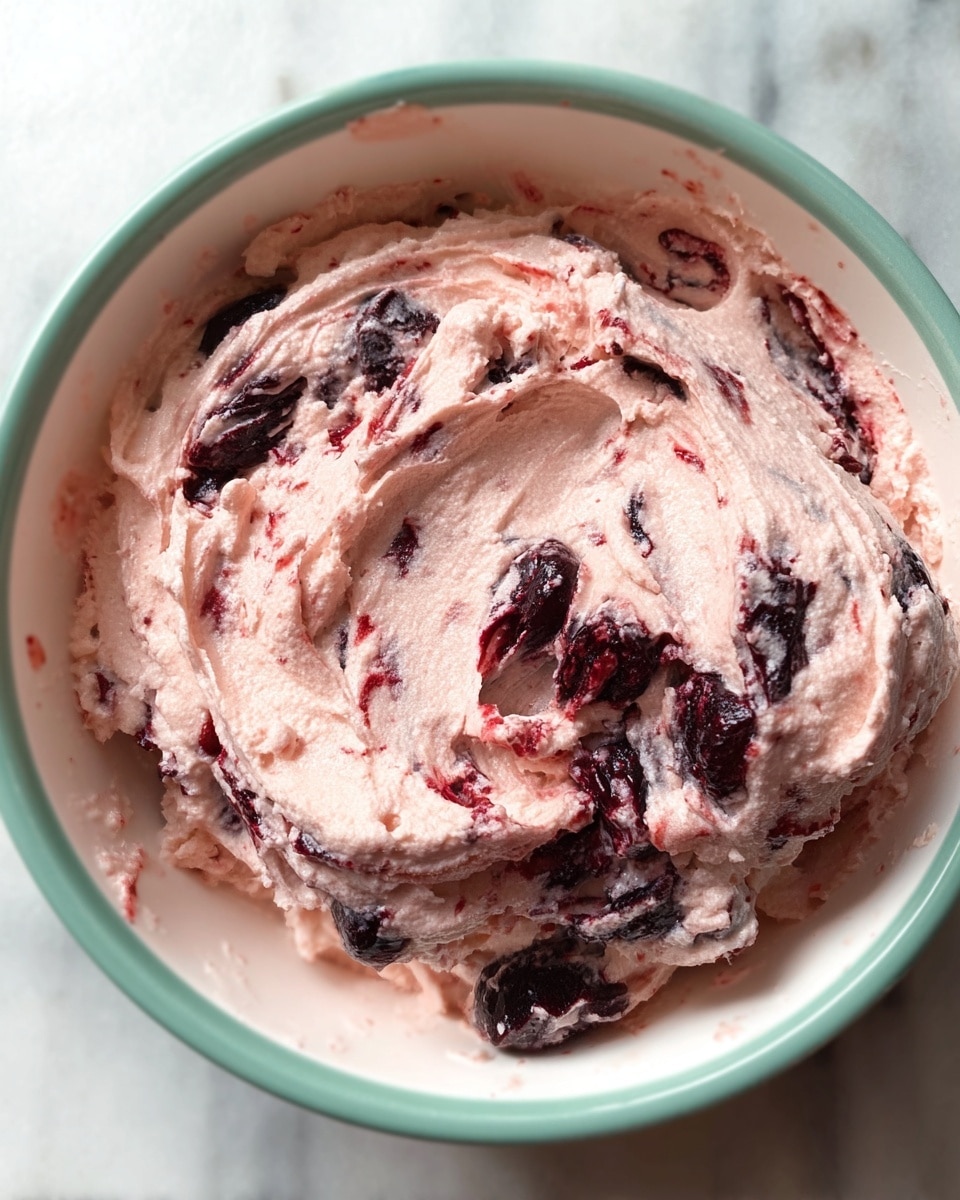 The image shows a close-up view of a creamy mixture swirled with dark purple and red fruit pieces inside a white bowl with a soft green rim. The mixture has a light pink color with thick, smooth, and fluffy texture, and the dark fruit pieces are unevenly spread throughout, creating a marbled effect. The bowl is placed on a white marbled surface. photo taken with an iphone --ar 4:5 --v 7
