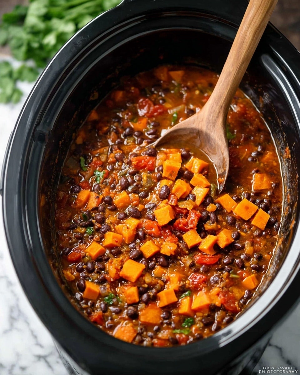 A black slow cooker is filled with a thick stew made of small black beans, bright orange sweet potato cubes, and diced red tomatoes, all mixed in a rich, reddish-brown sauce with visible small green and translucent bits from vegetables and herbs. A wooden spoon with a long handle is resting inside the cooker, partially submerged in the stew, showing the texture and mix closely. The background shows a blurred green leafy garnish on a white marbled surface. Photo taken with an iphone --ar 4:5 --v 7
