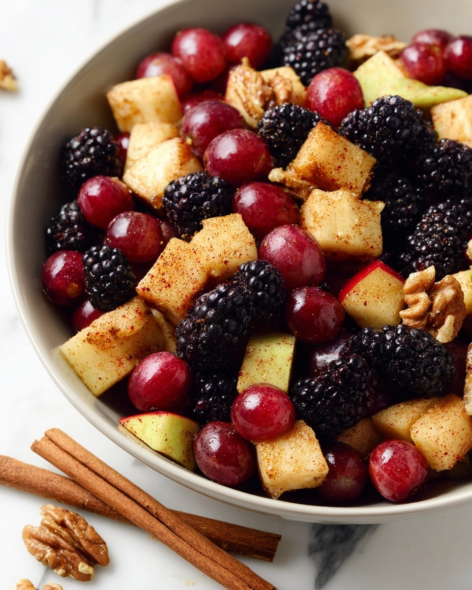 A close-up view of a fruit salad in a white bowl, showing three main layers: red grapes scattered throughout with a shiny, smooth texture; blackberries with a deep purple-black, bumpy surface filling most spaces between the fruits; and cubed apple pieces coated in a cinnamon-like spice, pale yellow with specks of red from the skin, all mixed evenly with walnut pieces adding a rough, brown layer. Two cinnamon sticks lie beside the bowl on a white marbled surface, enhancing the warm and fresh look. Photo taken with an iphone --ar 4:5 --v 7