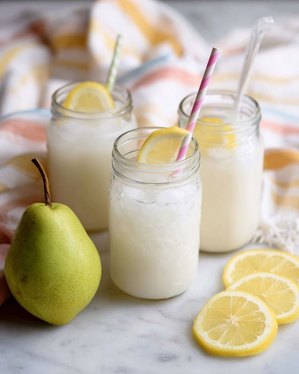 The image shows three clear glass jars filled with a creamy white drink that looks thick and smooth, each with ice cubes and a slice of lemon inside near the top. The jar in the front right has a pink and white striped straw, while the jar on the left has a white straw. A fresh green pear sits near the center on a soft white cloth with light yellow and peach stripes. To the bottom right, there are three lemon slices arranged together on a white marbled surface. The background is softly blurred, highlighting the refreshing drinks. photo taken with an iphone --ar 4:5 --v 7