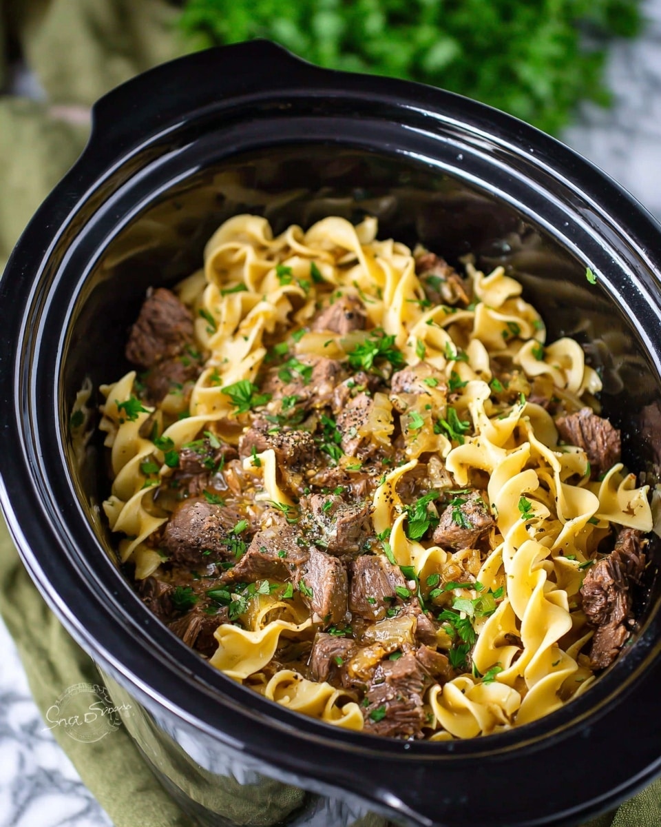 A close-up view of a black slow cooker filled with a layered beef and egg noodle dish; the bottom layer is tender chunks of brown beef mixed with some sauce and small bits of cooked onions, scattered throughout are wide, yellowish egg noodles folded and curled, topped with chopped fresh green parsley and sprinkled with black pepper. The slow cooker is set on a green cloth on a white marbled surface with blurred green herbs in the background. Photo taken with an iphone --ar 4:5 --v 7