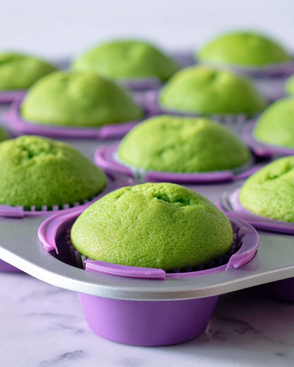 The image shows a close-up of several bright green muffins inside individual purple silicone molds which have small handles on each side, all placed on a silver metal muffin tray. The muffins have a soft, slightly cracked texture on top with a smooth, spongy look. The surface beneath the tray is a white marbled texture, providing a clean and light background that highlights the vivid green color of the muffins and the purple molds. Photo taken with an iphone --ar 4:5 --v 7