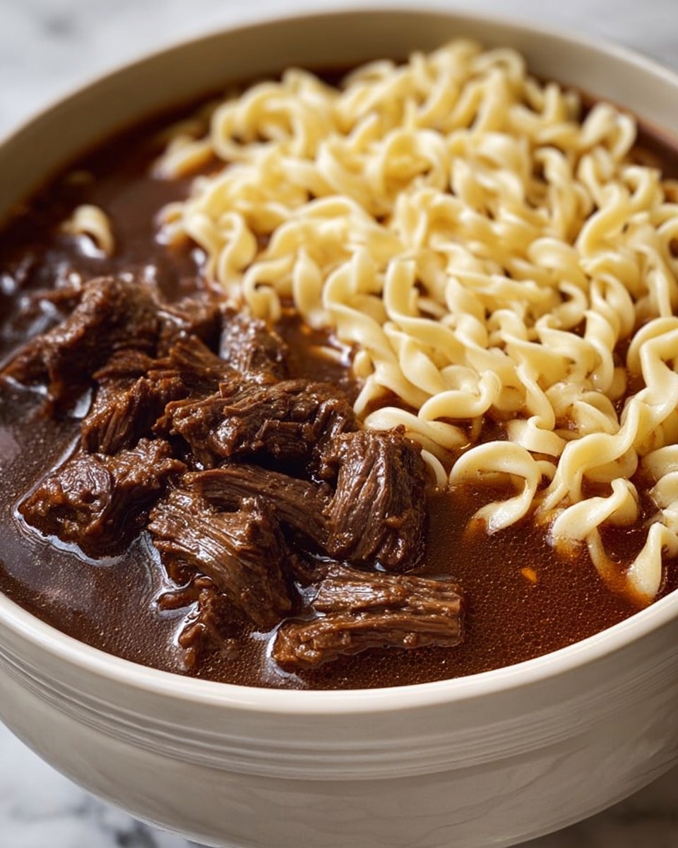 A close-up image of a white bowl filled with two main layers: the bottom layer contains a dark brown broth with visible chunks of tender, shredded beef scattered on top, showing a rich, glossy texture; the top layer is a bed of pale yellow egg noodles arranged neatly, occupying almost half of the bowl’s surface. The bowl sits on a white marbled surface, highlighting the contrast of colors and textures between the soft, light noodles and the thick, meaty broth. photo taken with an iphone --ar 4:5 --v 7