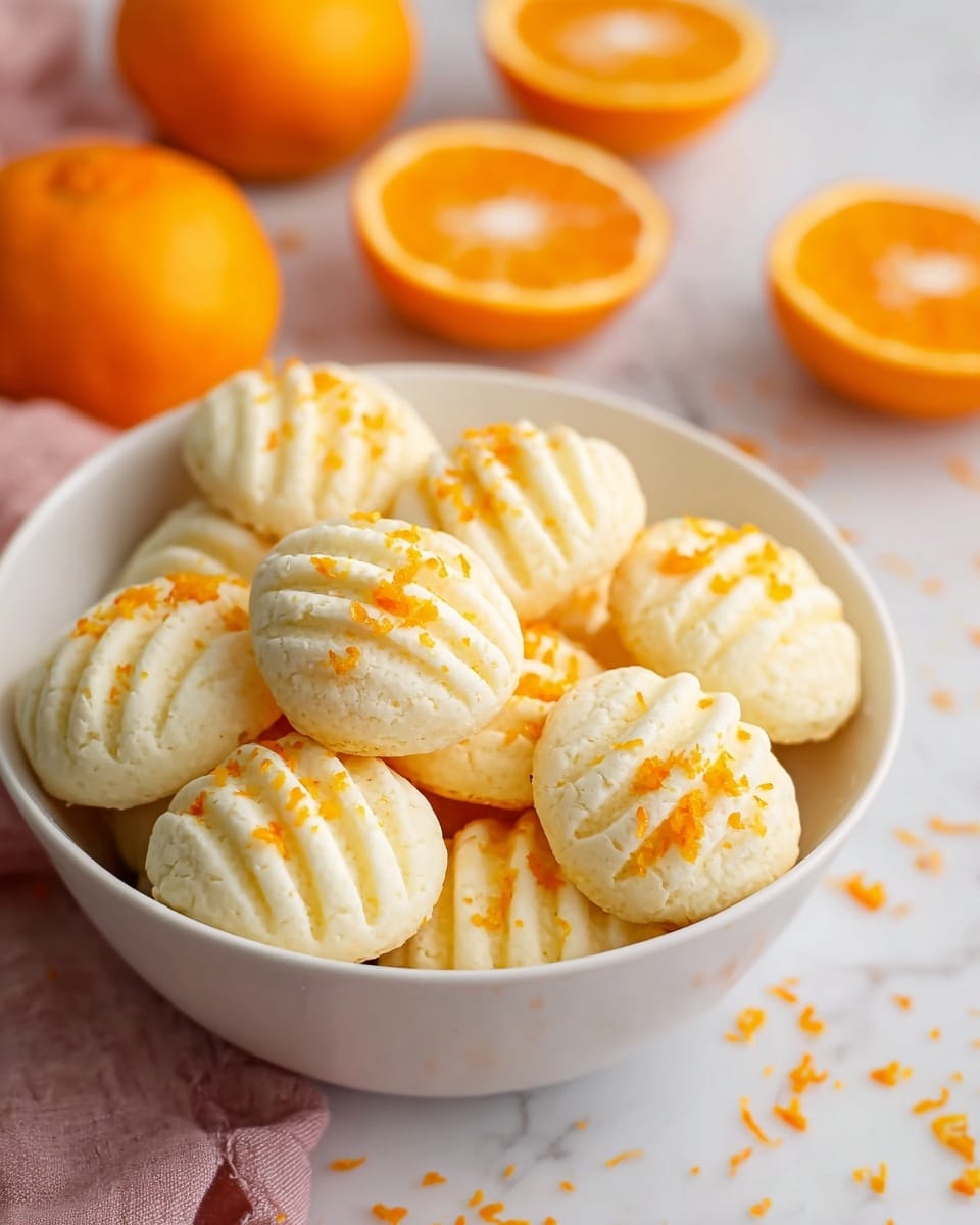 A white bowl filled with small, round, white cookies that have ridges running across the top surface in a curved pattern; each cookie is sprinkled with small bright orange zest pieces. The bowl is placed on a white marbled surface with some orange zest scattered around it, and four halved oranges sit in the background out of focus. A soft pink cloth is partially visible underneath the bowl on the left side. photo taken with an iphone --ar 4:5 --v 7
