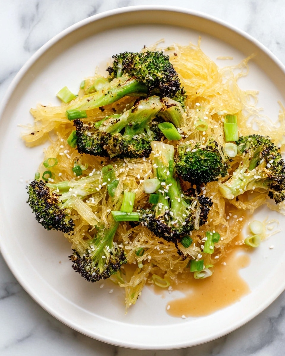 A white plate holds a serving of cooked spaghetti squash with several pieces of roasted broccoli on top. The spaghetti squash strands are pale yellow and look soft with a slightly shiny texture from a light sauce. Bright green broccoli florets are charred at the edges, giving a mix of dark green and brown colors. Thin slices of light green scallions are scattered among the vegetables, along with white sesame seeds sprinkled on the top. A light brown sauce pools around the edges of the plate on a white marbled surface. Photo taken with an iphone --ar 4:5 --v 7