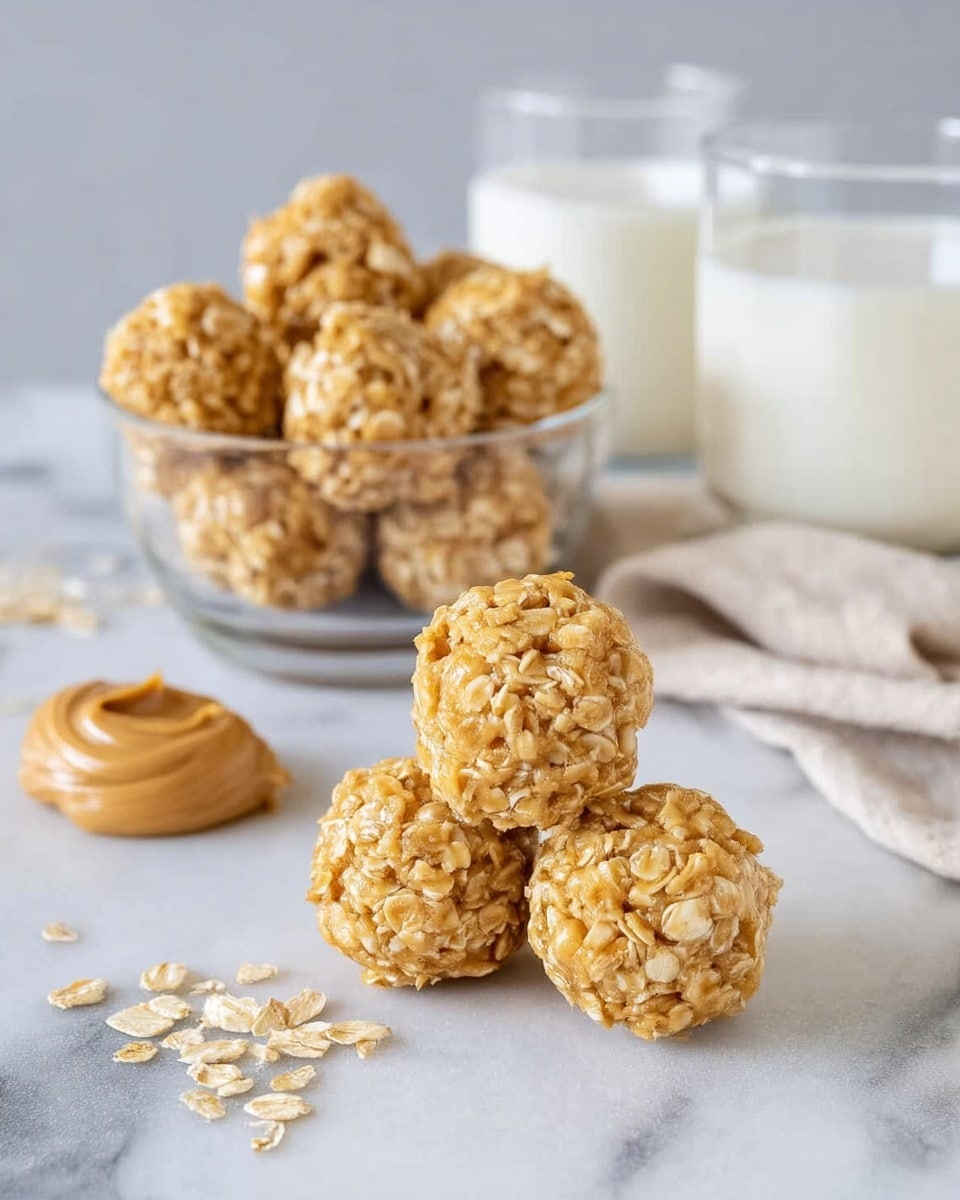 A close-up of three small peanut butter oat balls stacked in a pyramid on a white marbled surface, showing a rough texture with visible oats and a golden brown color. Behind the pyramid is a clear glass bowl filled with many more of the same oat balls. To the left, a dollop of peanut butter lies on the surface with some scattered oats nearby. On each side behind the bowl, there are two clear glass cups filled with white milk, and a light-colored cloth is placed near the cup on the right. The background is soft and neutral, keeping all focus on the snack and milk. photo taken with an iphone --ar 4:5 --v 7
