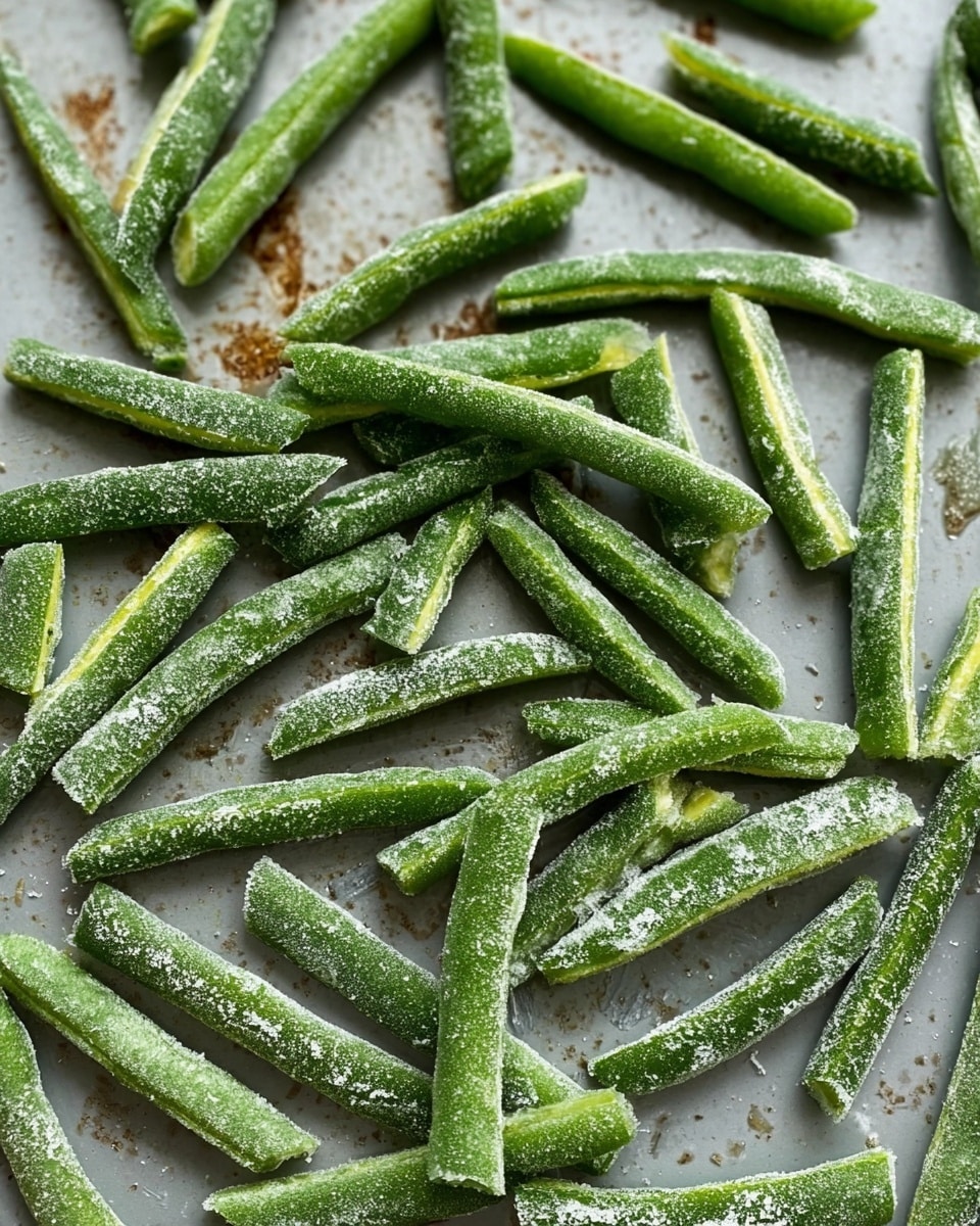 The image shows many cut green beans spread out in a single layer on a flat gray surface with some light brown marks. Each green bean is bright green and slightly frosted with a white powdery coating, giving them a textured look. The beans vary in length, some whole and some split, with clear details of their edges and slight curves. The background is a simple flat surface with a few dark and light spots, making the green beans the main focus. Photo taken with an iphone --ar 4:5 --v 7