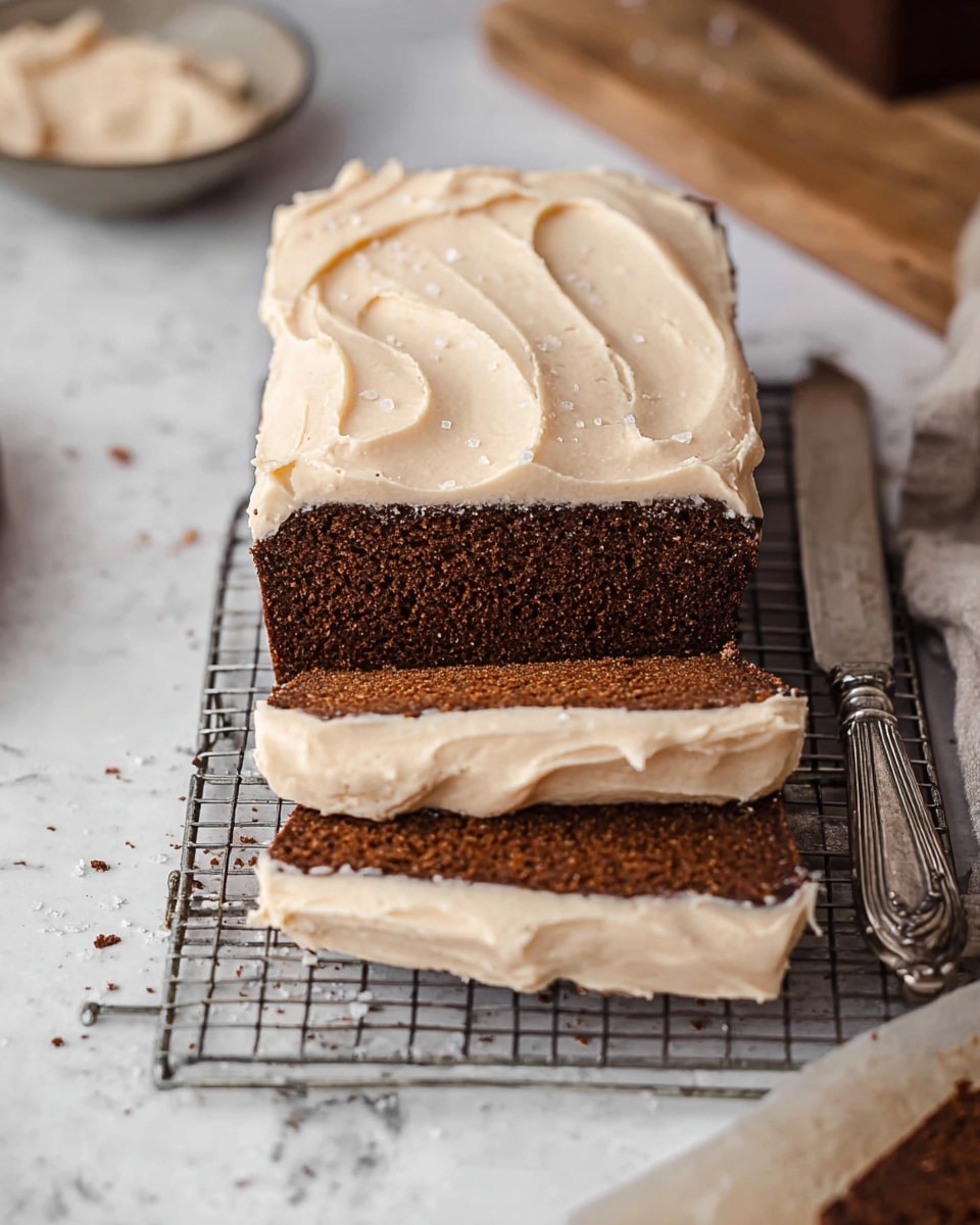 The image shows a loaf cake sliced into thick pieces, placed on a metal cooling rack over a white marbled texture. The cake has two visible layers: the bottom layer is a dense, dark brown spongy texture, and the top layer is a thick, creamy light beige frosting spread evenly over the cake, with soft swirls and tiny bits visible in its texture. A silver knife with a detailed handle lies beside the cake, and small crumbs and a few sprinkles of coarse salt are scattered around, adding to the rustic feel. In the background, there are blurred elements including a wooden board and some more frosting on parchment paper. photo taken with an iphone --ar 4:5 --v 7