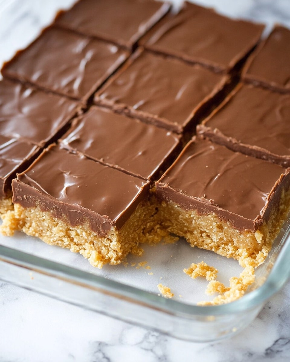 The image shows a close-up of a dessert with two layers inside a glass baking dish sitting on a white marbled surface. The bottom layer is thick, crumbly, and light golden brown with a slightly rough texture, like a cookie or oat base. The top layer is smooth and creamy chocolate, medium brown in color, spread evenly and cut into neat square pieces. One square piece is missing, revealing the clean cut between the layers with some crumbs around the empty space. Photo taken with an iphone --ar 4:5 --v 7