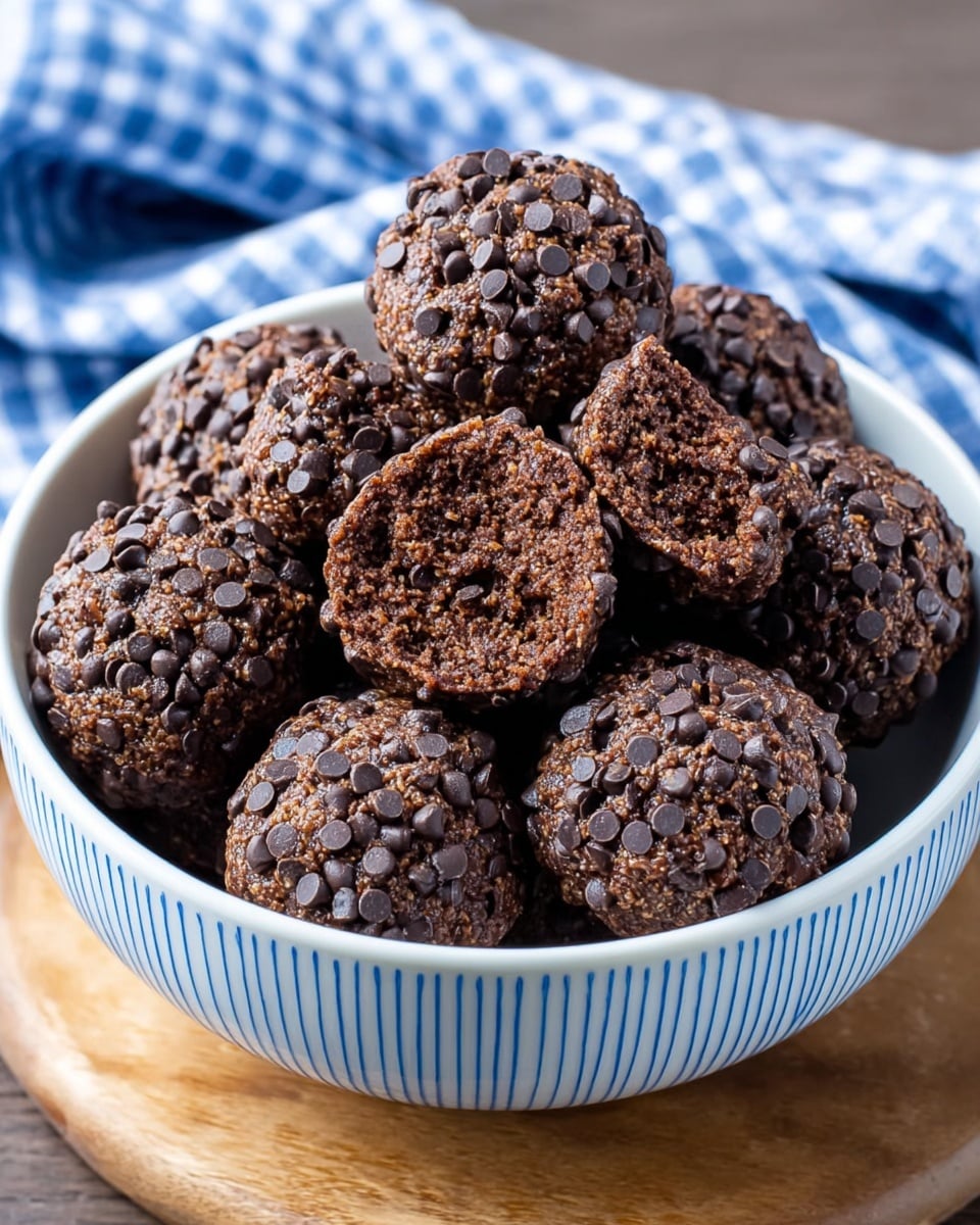 A white bowl with thin blue stripes holds round chocolate treats covered fully in small, dark chocolate chips. There are about a dozen pieces inside the bowl, piled closely. Each treat has a rough, textured surface from the chocolate chips, giving a bumpy look. One piece near the front is bitten, showing a dense, dark brown inside that looks moist and crumbly. The bowl sits on a wooden board with a blue and white checkered cloth in the background, but the main focus is on the chocolate treats. photo taken with an iphone --ar 4:5 --v 7