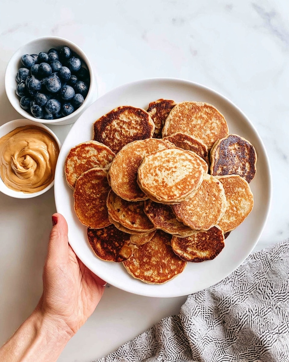 A white plate filled with a layered stack of about twenty small, round pancakes, each showing a golden brown to darker toasted color with slightly uneven edges and a soft, porous texture. The plate is held by a woman's hand from below, resting against a white marbled surface. To the upper left, two small white bowls appear: one filled with fresh, plump blueberries, dark blue to almost black in color, and the other containing a swirl of light brown peanut butter with a creamy, slightly chunky texture. A patterned grey and white cloth is seen on the right side of the image. photo taken with an iphone --ar 4:5 --v 7