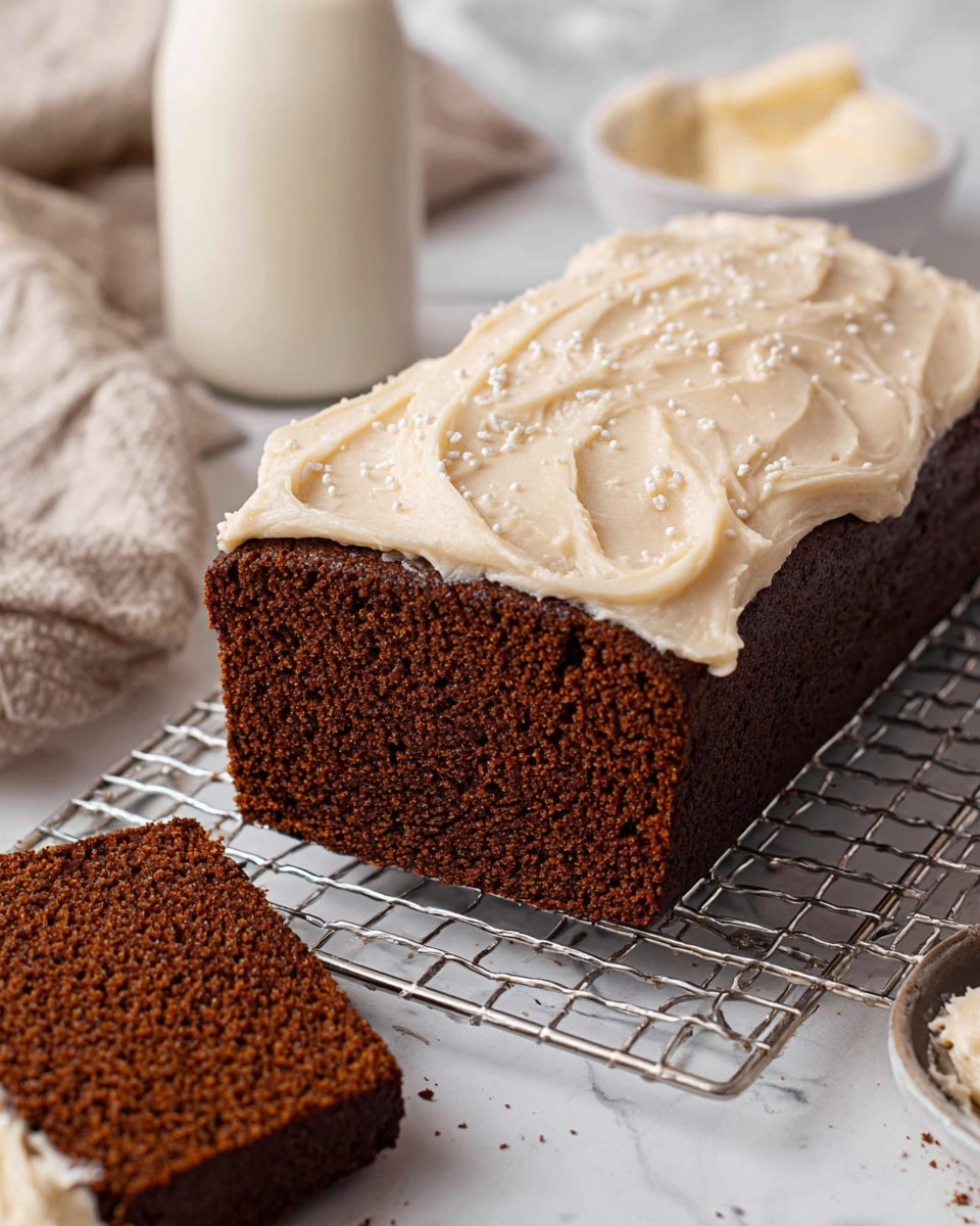 A rectangular dark brown loaf cake with a rough, moist texture is placed on a metal cooling rack over white marbled surface. The cake is topped with one thick layer of light beige creamy frosting, spread unevenly with swirled patterns, and sprinkled lightly with small white flakes. A slice of the same cake is cut and placed in front, showing the soft crumb inside. In the blurred background, there is a tall glass of milk and a white bowl with more frosting, all on the white marbled surface photo taken with an iphone --ar 4:5 --v 7