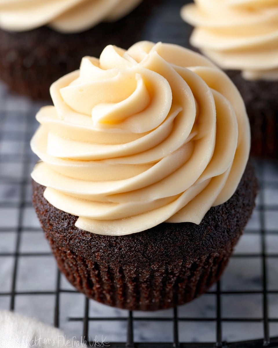 A close-up view of a chocolate cupcake topped with a large swirl of creamy, light beige frosting. The cupcake base is dark brown with a slightly rough texture, showing a moist crumb surface. The frosting is thick, smooth, and glossy, piped in wide, even spiral layers starting from the edge of the cupcake and rising in smooth curved peaks toward the center. The cupcake is set on a black cooling rack with a white marbled texture background partly visible beneath it. Photo taken with an iphone --ar 4:5 --v 7