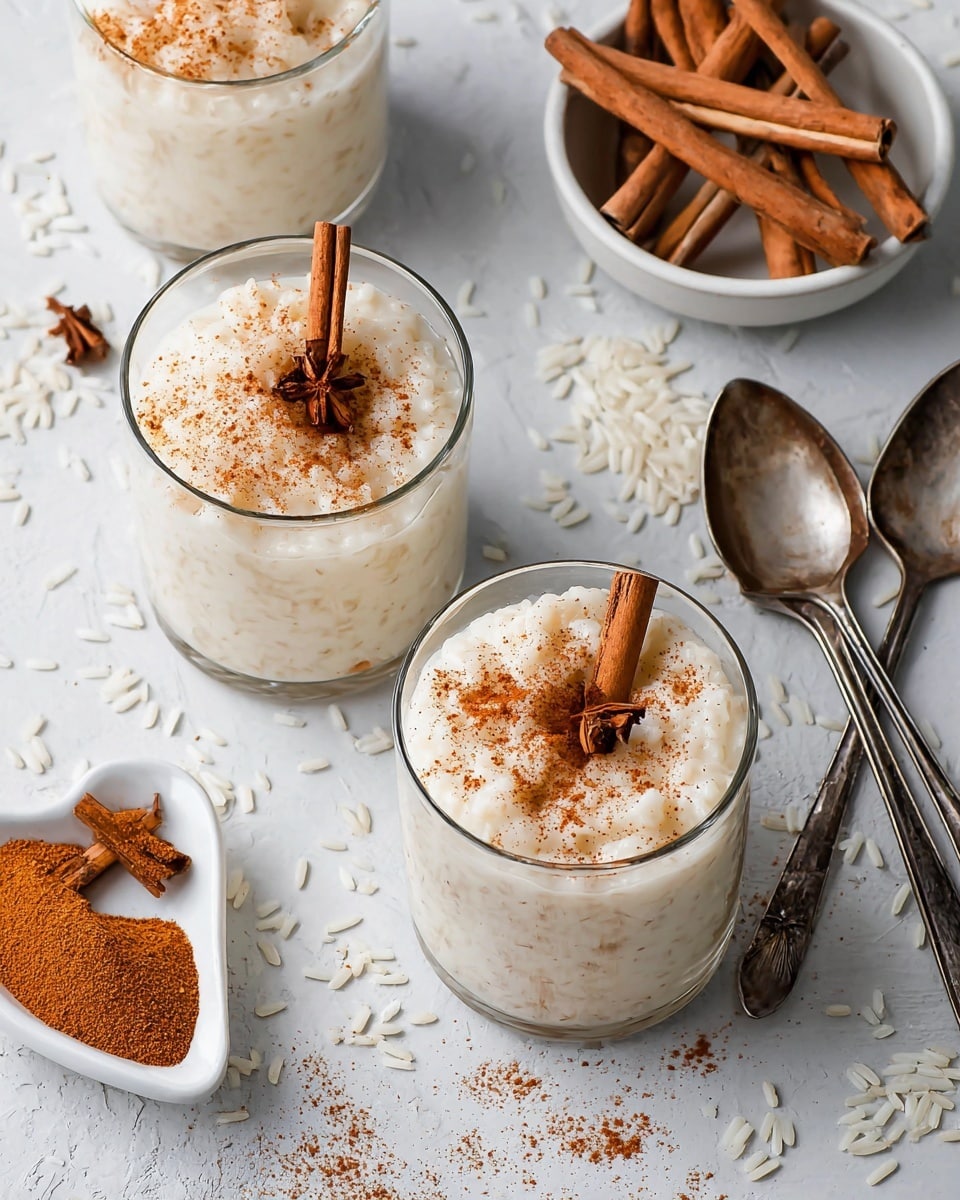 Three clear glass cups filled with creamy white rice pudding, each topped with a sprinkle of light brown cinnamon powder and a small brown cinnamon stick piece. The pudding has a soft, slightly lumpy texture with visible rice grains throughout. The cups are placed on a white marbled surface with scattered white rice grains around them. There is a small white bowl to the side filled with longer cinnamon sticks and a heart-shaped white dish holding ground cinnamon powder. Two vintage silver spoons lie on the surface next to the dishes. Photo taken with an iphone --ar 4:5 --v 7