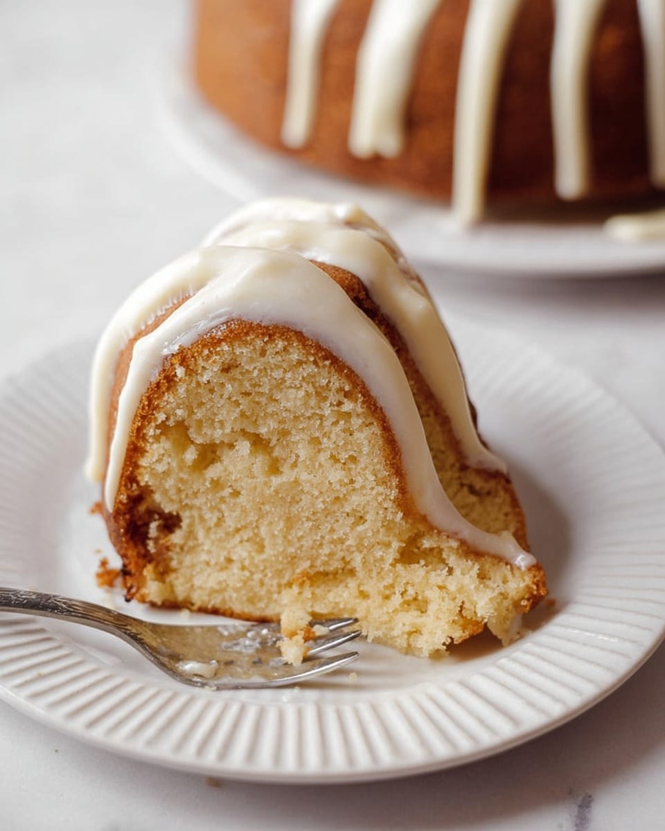 A single slice of light golden brown bundt cake sits on a white ridged plate with soft, thick white icing draped over the outer edge in flowing lines. The cake slice reveals a moist, fluffy inside with a few darker areas near the edges, showing a tender crumb texture. A silver fork with a piece of the cake is cut from the side, resting near the front of the slice. In the background, the rest of the cake is slightly out of focus on a white marbled surface. photo taken with an iphone --ar 4:5 --v 7