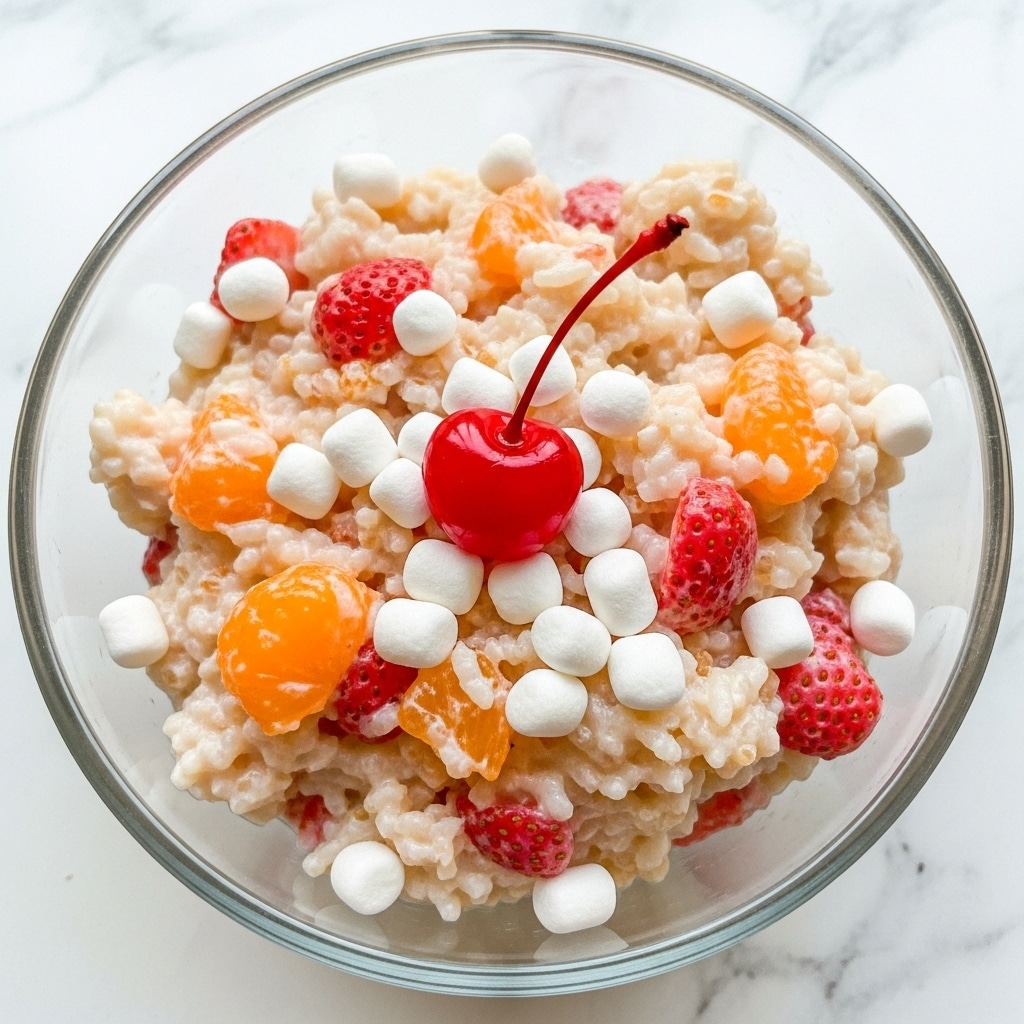 A clear glass bowl filled with a creamy, off-white mixture containing small pieces of light orange mandarin, pinkish-red strawberries, and white miniature marshmallows, topped with a bright red maraschino cherry with its stem in the center. The texture of the dish looks soft and fluffy with a mix of fruit chunks and marshmallows scattered throughout. The bowl is set on a white marbled surface. Photo taken with an iphone --ar 4:5 --v 7