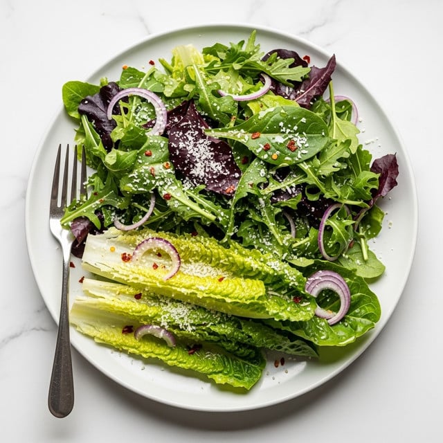 A white plate holds a fresh green salad with two main layers of leafy greens: a base layer of light green romaine lettuce with crisp texture and a top layer of darker green and purple-tinted leafy greens. Thin slices of light purple shallots peek through the leaves, and the salad is sprinkled all over with fine white grated cheese and tiny flakes of red chili pepper, adding texture and color contrast. A silver fork rests on the left side of the plate, partly touching the salad. The whole scene is set on a white marbled surface. photo taken with an iphone --ar 4:5 --v 7