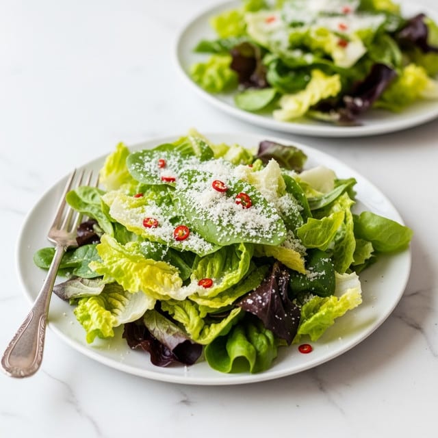 Two white plates with fresh Caesar salad are set on a white marbled surface. The front plate shows leafy greens in different shades of green, ranging from light to dark, forming the main layer with a loose, textured look. A light sprinkling of finely grated white cheese is scattered evenly on top, along with small red chili flakes for a touch of color. A silver fork lies on the front left side of the plate, its handle resting on the plate's edge. The second plate in the background holds a similar salad, slightly out of focus but showing the same mix of green leaves and cheese. Photo taken with an iphone --ar 4:5 --v 7