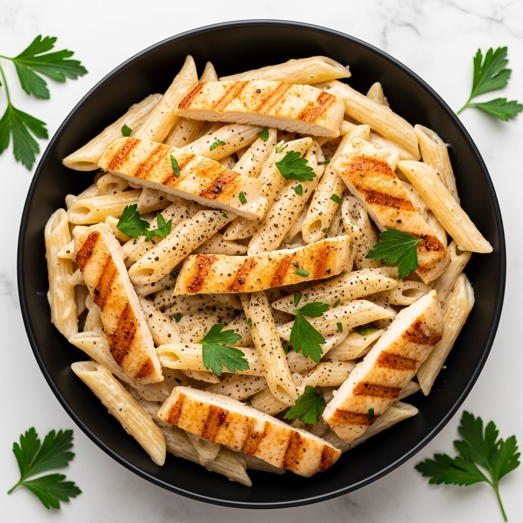 A black bowl filled with creamy penne pasta and grilled chicken pieces mixed together. The pasta is coated in a light beige creamy sauce, with golden-brown grilled chicken strips scattered evenly throughout. Small pieces of green parsley are sprinkled on top along with black pepper specks, adding color contrast. The bowl sits on a white marbled surface with some parsley leaves around it. Photo taken with an iphone --ar 4:5 --v 7