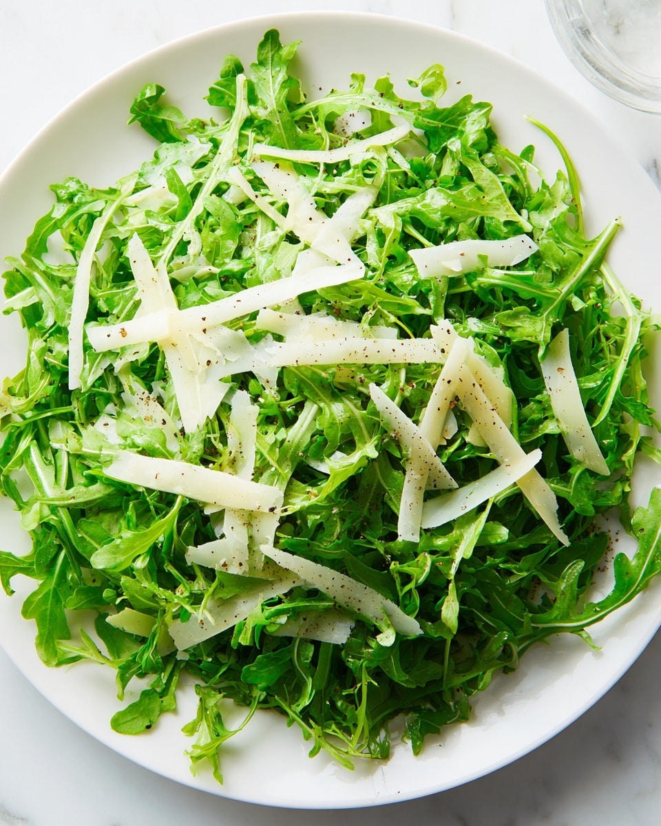 A white plate filled with a fresh arugula salad showing two main layers; the base layer is made of bright green arugula leaves with a slightly crinkled texture, scattered evenly across the plate, and the top layer consists of thin, long, pale yellow cheese shavings spread lightly over the greens, with visible speckles of black pepper. The salad sits on a white marbled surface, and there is a soft natural light illuminating the vibrant colors and fresh textures. photo taken with an iphone --ar 4:5 --v 7