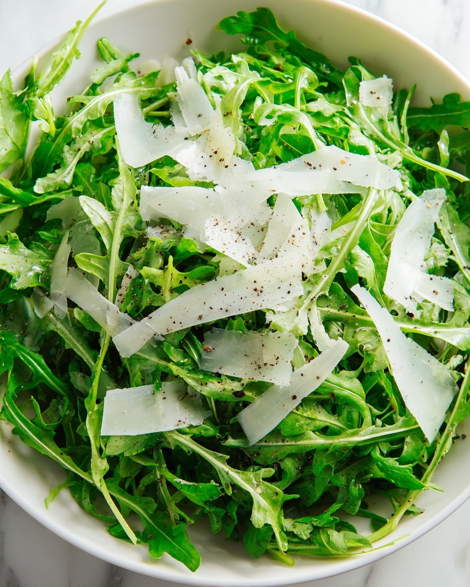 A close-up view of a fresh arugula salad in a white bowl, showing two main layers: the bottom layer of bright green arugula leaves with a slightly jagged texture, and the top layer of thin, translucent white cheese shavings scattered unevenly across the arugula. The salad is lightly sprinkled with black pepper and visible drops of olive oil, giving a glossy appearance and slight shine to parts of the leaves. The bowl sits on a white marbled surface. photo taken with an iphone --ar 4:5 --v 7